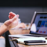 Hand, Laptop, Smartphone und Notizbuch auf Tisch bei Arbeitssituation.