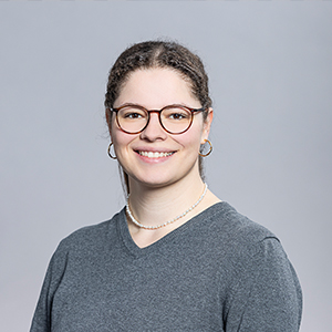 Lächelnde junge Frau mit Brille, grauem Pullover und Perlenkette vor grauem Hintergrund.