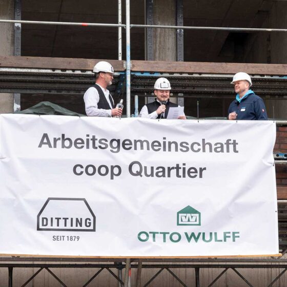 Drei Bauarbeiter mit Helmen bei einer Baustellenzeremonie mit Banner "Arbeitsgemeinschaft coop Quartier".