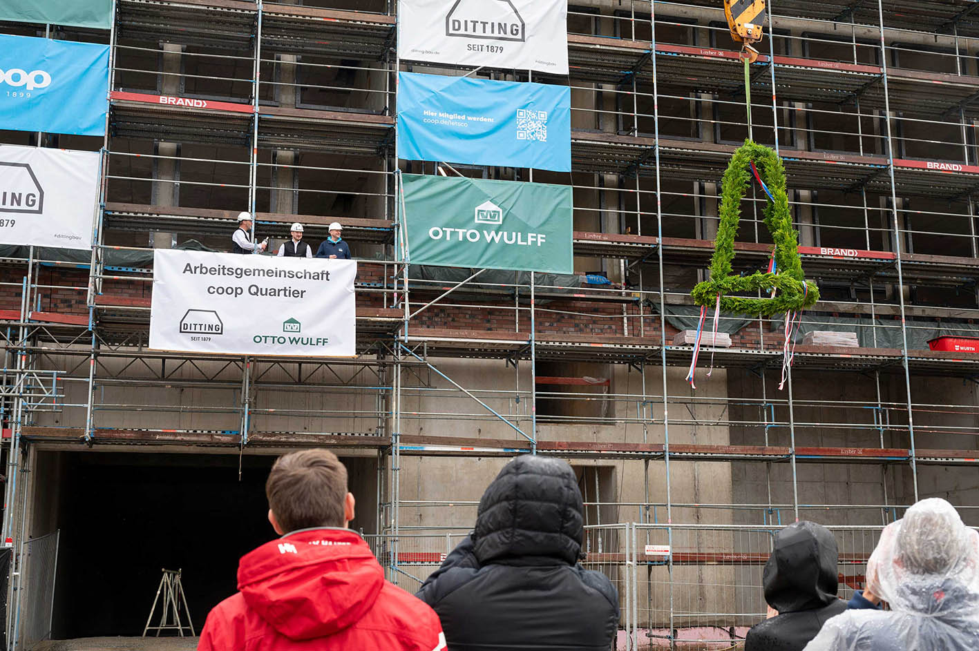Bauarbeiten an einem Mehrfamilienhaus mit Feier und Grußbann, Menschen im Regen.