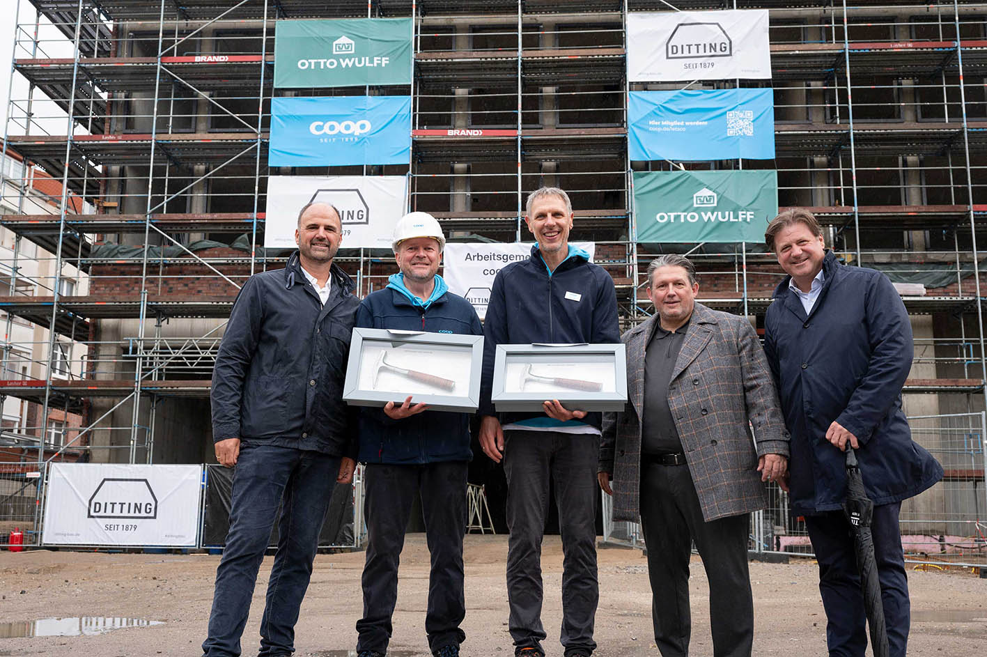 Bauarbeiter und Manager bei Baustellenübergabe vor Gerüst und Bauplänen.