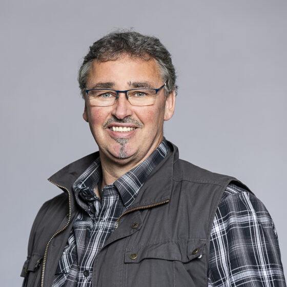 Porträt eines lächelnden Mannes mit Brille, kariertem Hemd und dunkler Weste vor grauem Hintergrund.