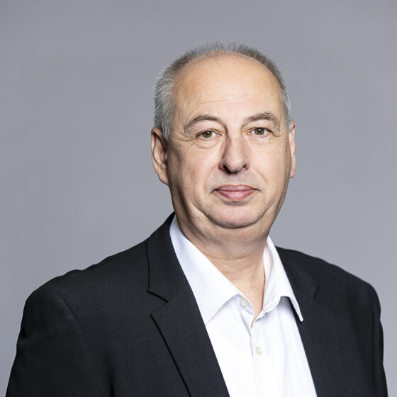 Porträt eines Mannes in formeller Kleidung vor grauem Hintergrund.