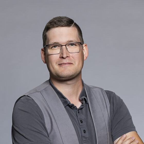 Selbstbewusster Mann mit Brille in grauem Hemd und dunkler Weste vor grauem Hintergrund.