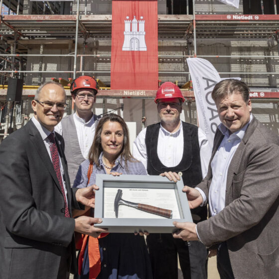 Gruppenfoto bei Bauprojekt-Feier mit symbolischem Hammer und Bauhelm, im Hintergrund Gerüst.