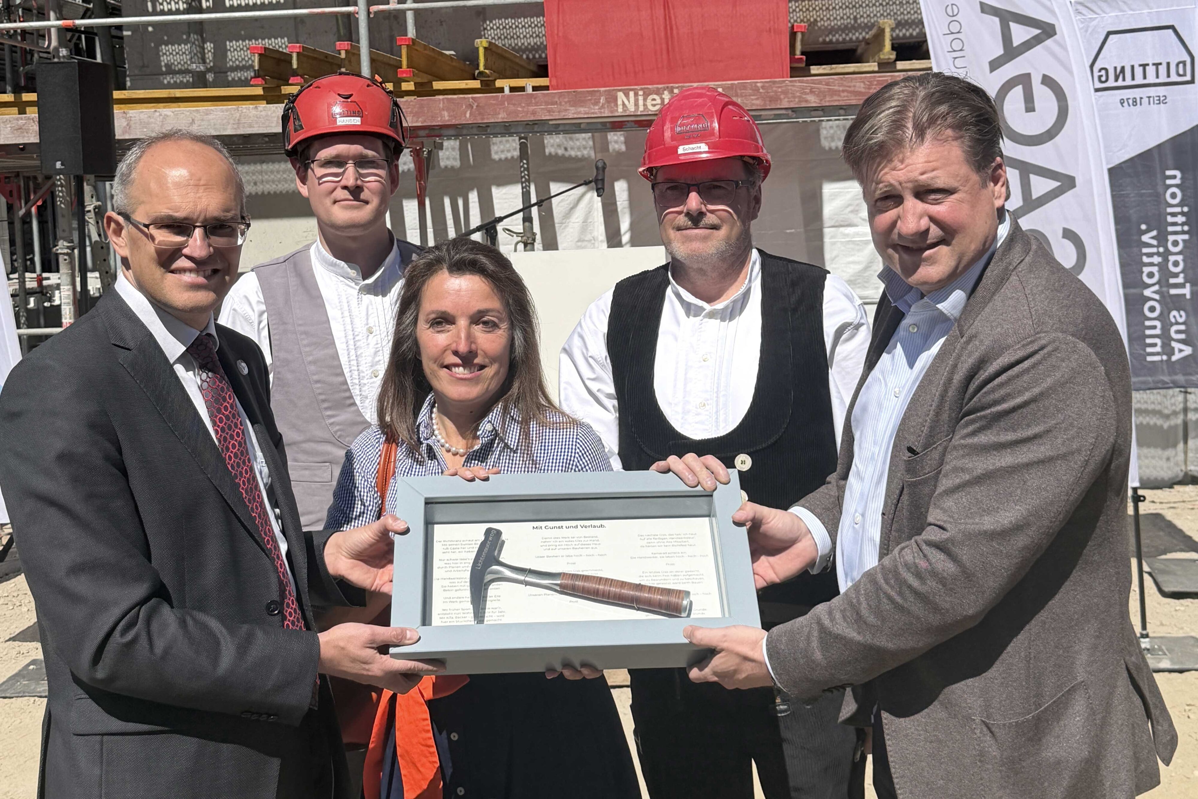 Gruppenfoto bei Bauprojektübergabe mit Männern, Frauen, Helm, Zertifikat und Baustelle im Hintergrund.