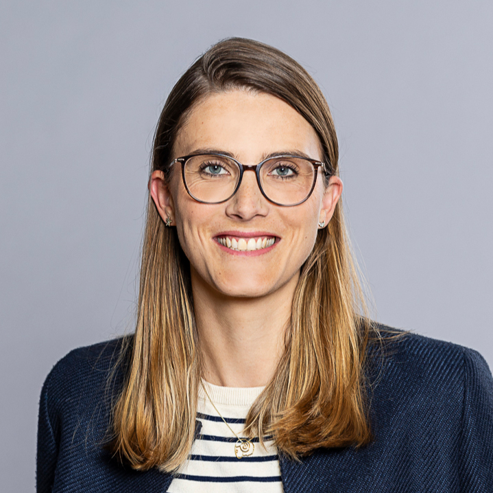 Porträt einer professionellen, lächelnden Frau mit Brille vor grauem Hintergrund.