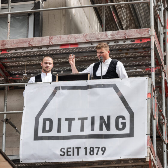 Zwei Männer beim öffentlichen Vortrag auf einer Baustellenbühne mit "Ditting" Banner.