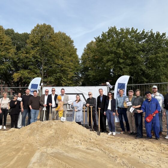 Gruppenfoto bei Baustart mit Menschen vor Bauzaun und Baufahrzeugen in urbaner Umgebung.